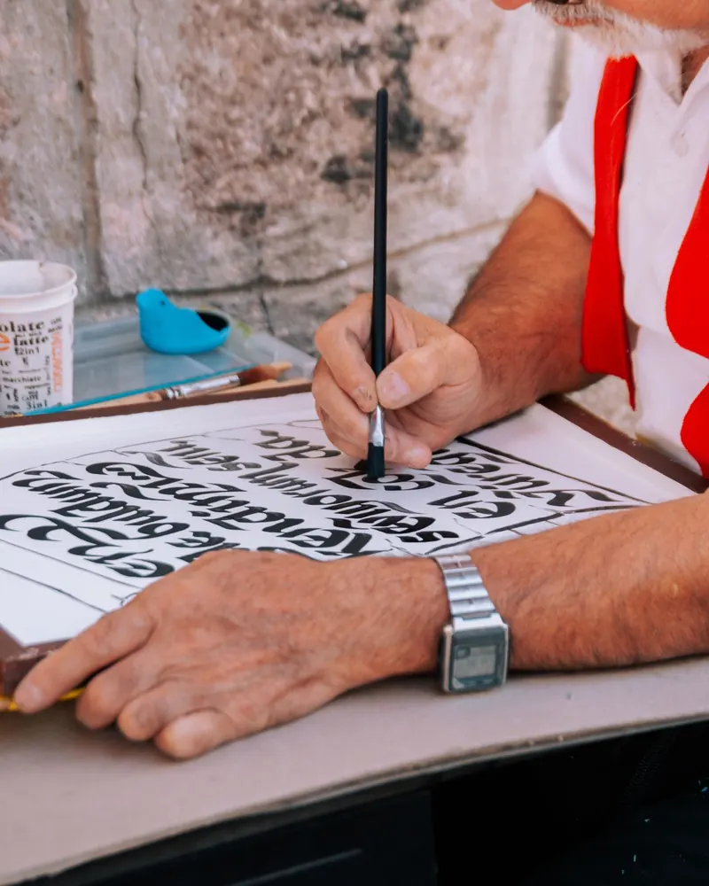 Mão a segurar um pincel fino na vertical sobre uma folha com letras pintadas a preto, enquanto a outra mão fixa o papel na bancada