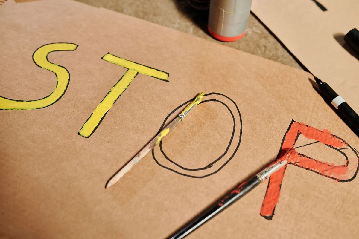 Letras contornadas a preto sobre cartão, com a primeira parte já preenchida a amarelo e a última a vermelho, e dois pincéis pousados sobre o trabalho
